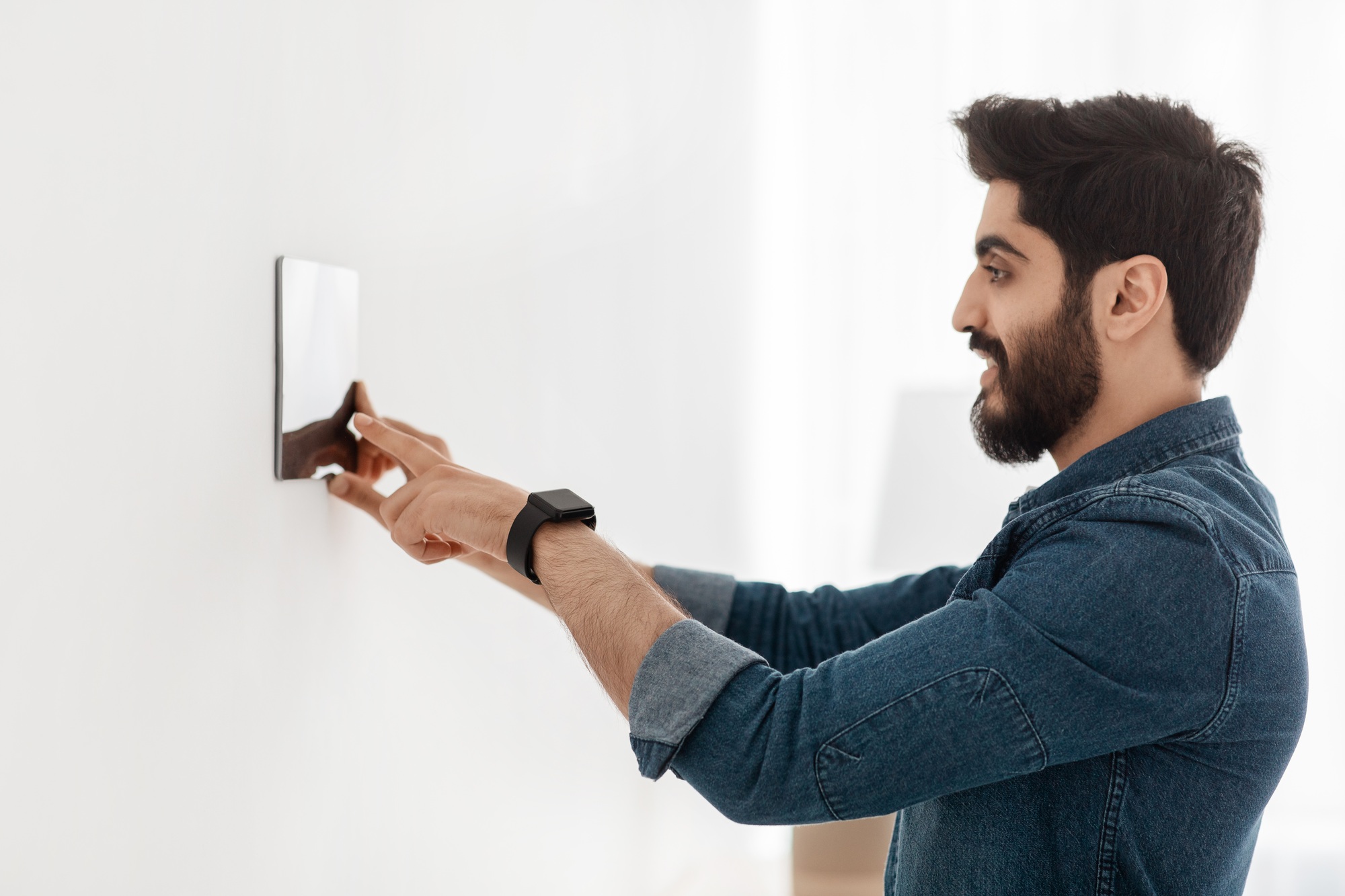 Bearded man controlling house with digital touch screen panel installed on light wall in the living